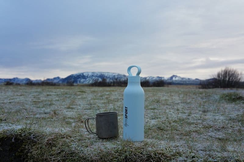 An insulated bottle and an enamel mug on frosted grass, mountains in the distance.