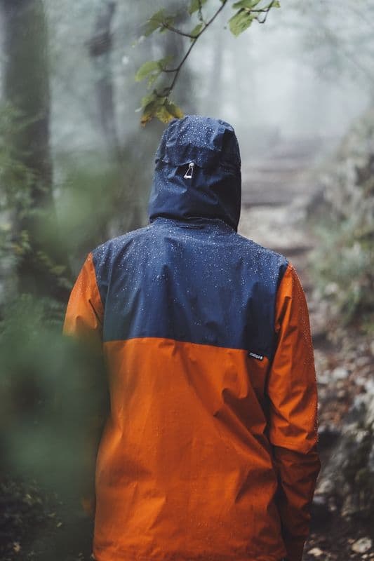 The back of a hooded orange and navy waterproof shell in wet forest.