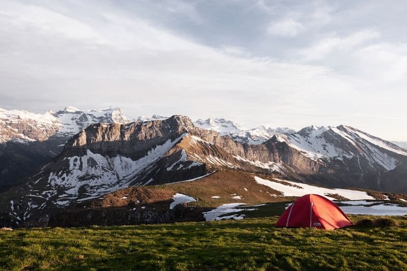 A red dome tent on a green slope below a snow-covered mountain range.
